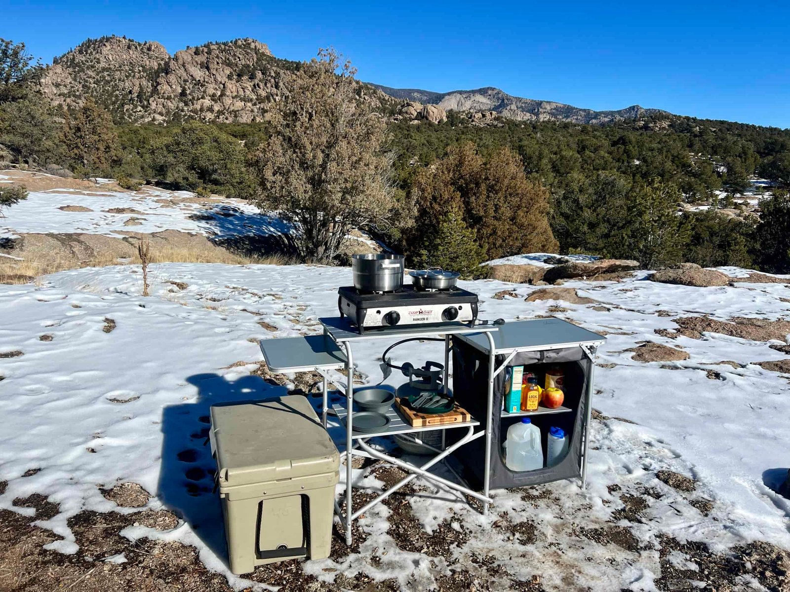 The Mountain Summit Gear Deluxe Roll Top Kitchen stands on snowy ground with rocky mountains and trees in the background. The table is loaded with a two-burner stove, cookware, and food items. A cooler is sitting next to it.