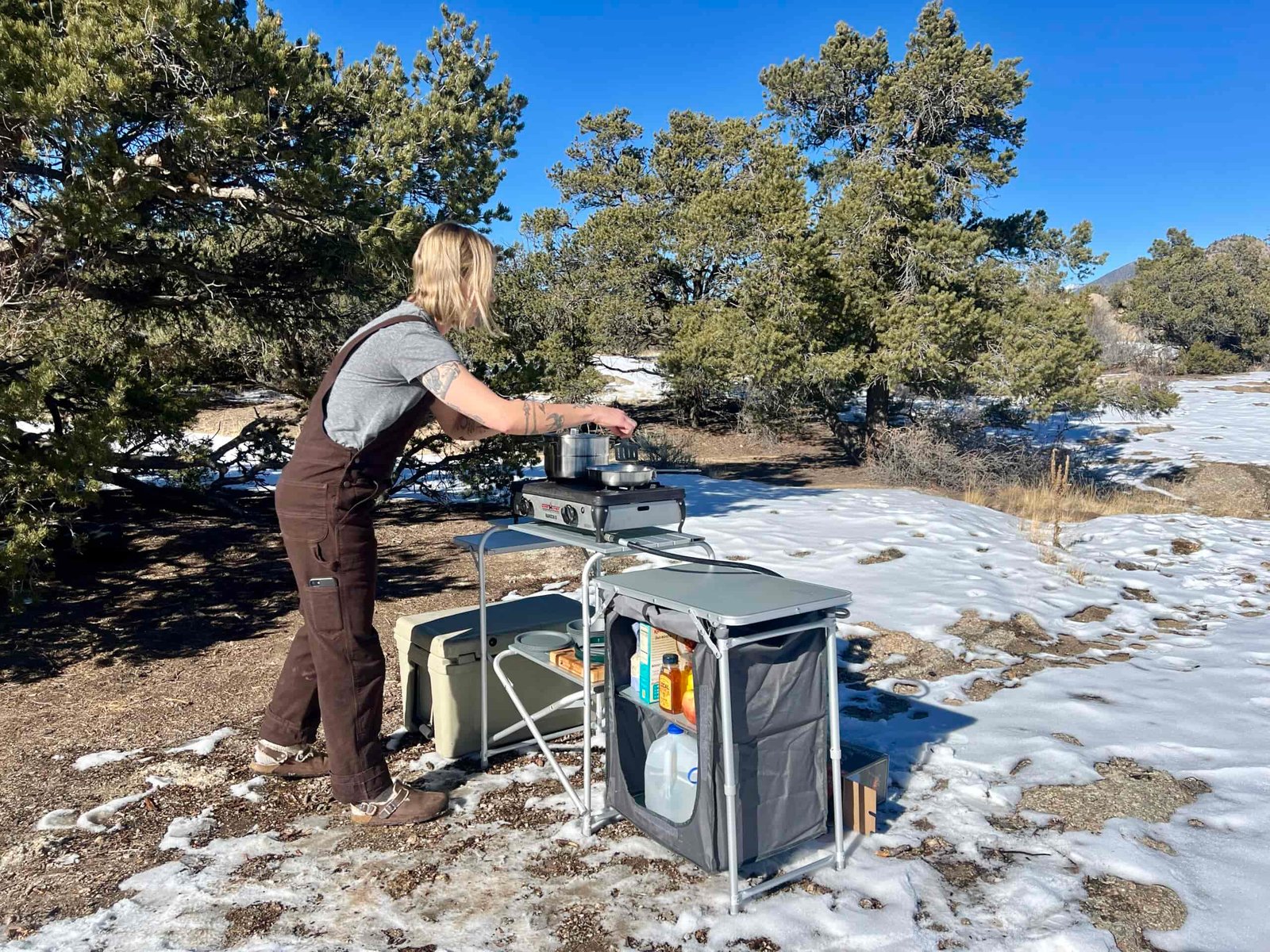 The Mountain Summit Gear Deluxe Roll Top Kitchen stands on snowy ground with oak trees behind it. A person in brown overalls and a gray t-shirt stands at the table cooking.