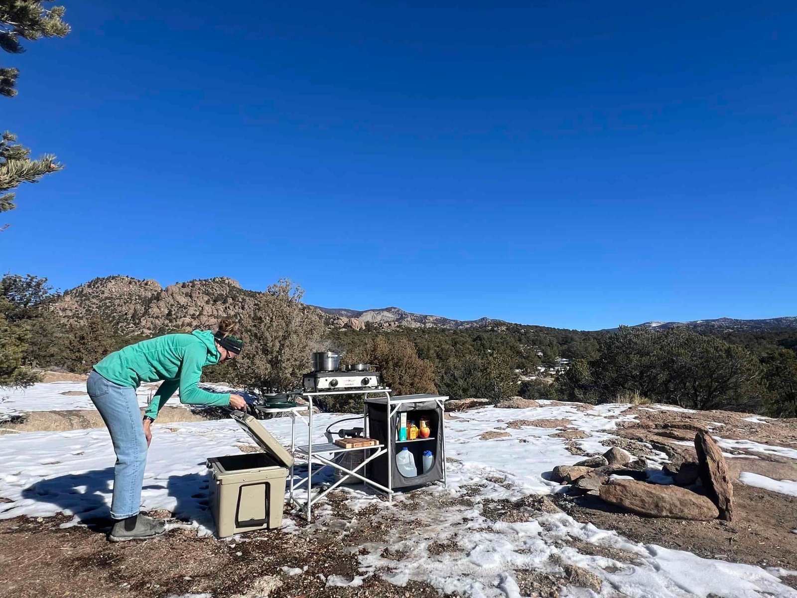 The Mountain Summit Gear Heavy Duty Roll Top Kitchen stands in snow and dirt with a backdrop of rocky hills and oak trees. A person standing next to the table wearing jeans, and a teal sweater, is looking into an ice chest.