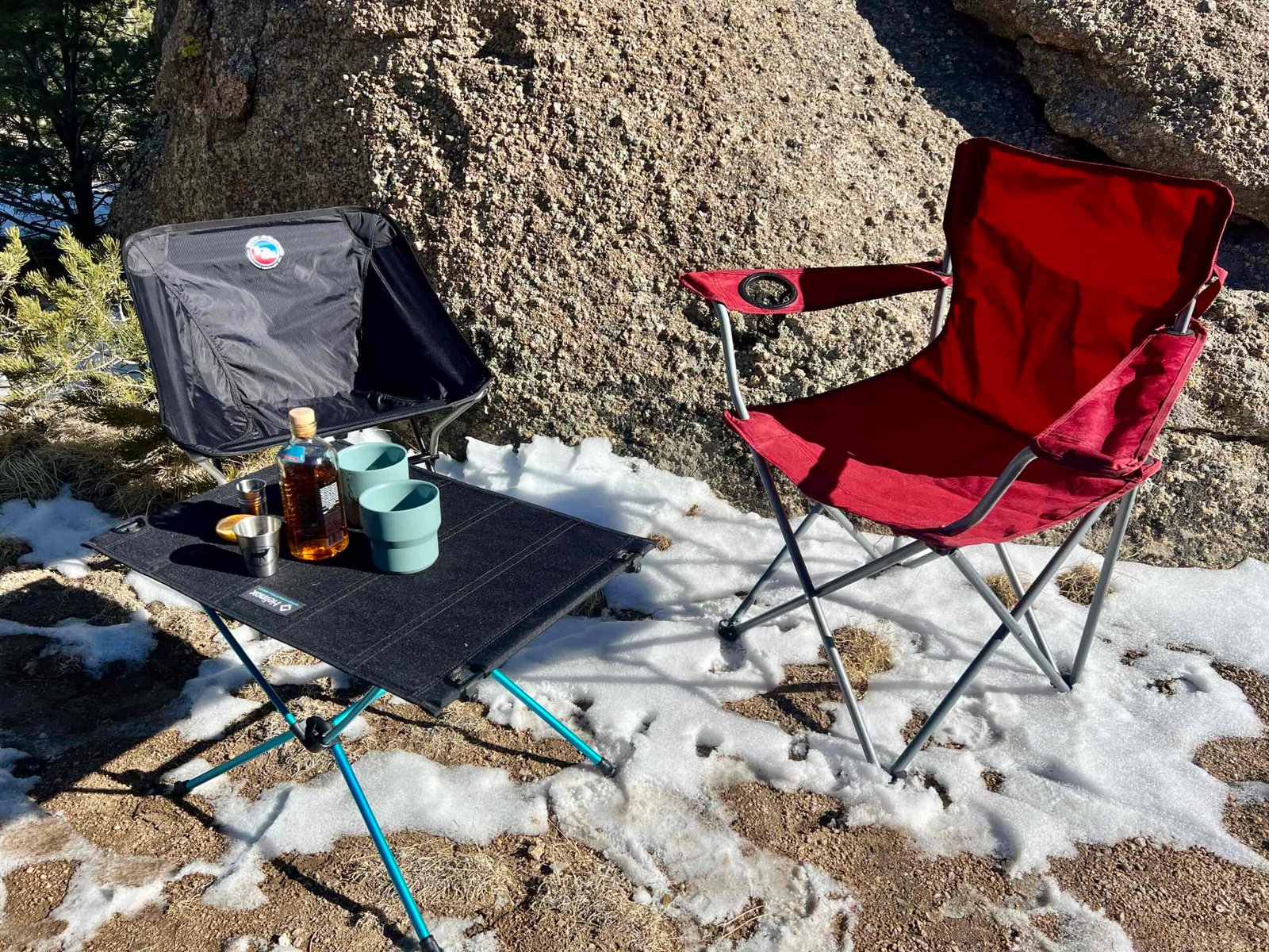 The Helinox Table One Hard Top sits on dirt and snow with cups and a bottle of whiskey on top. Empty red and black chairs sit around the table. A large boulder is in the background.