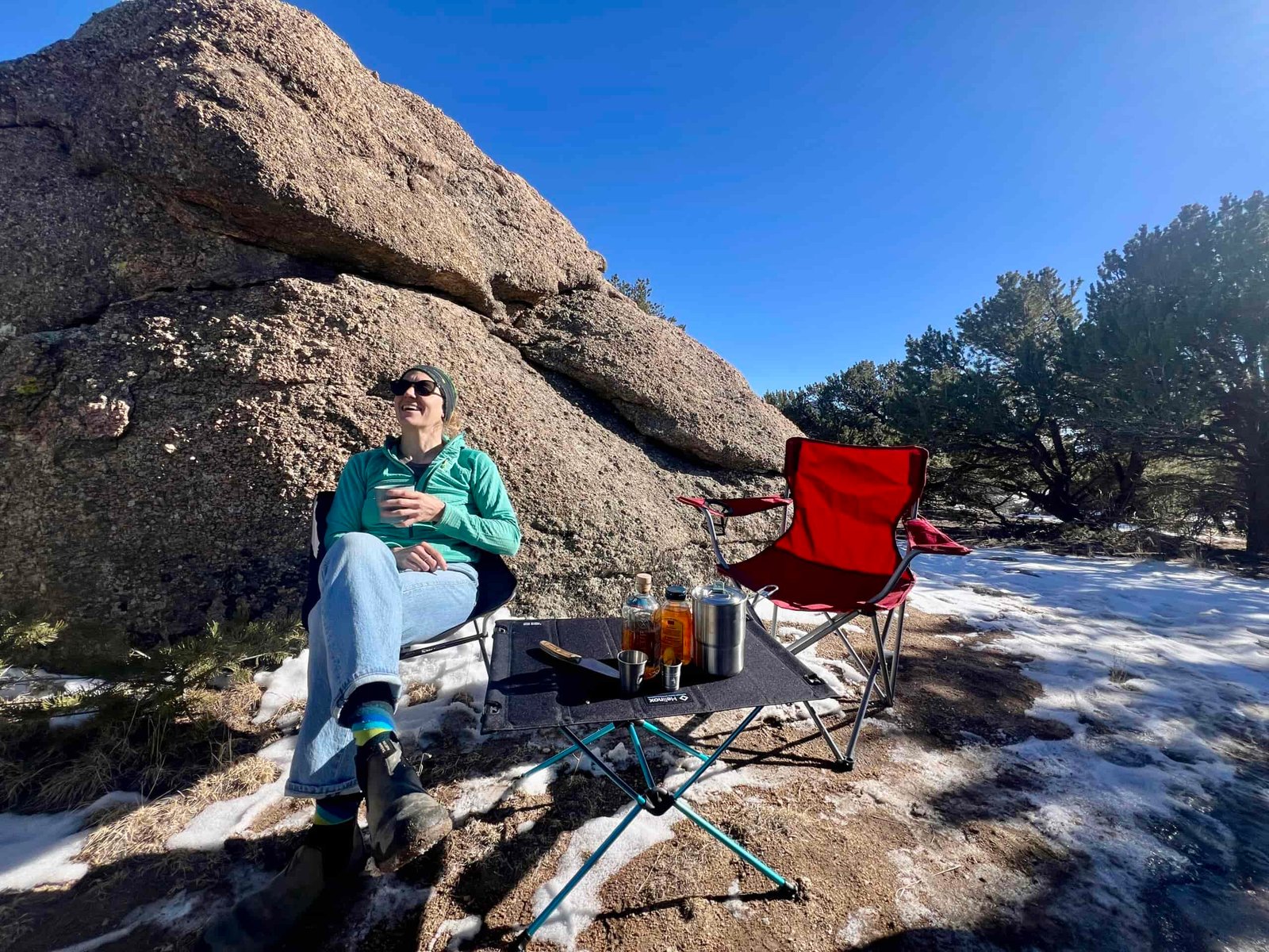 The Helinox Table One Hard Top has beverages on it. A person is smiling and looking ahead with a teal sweater, sunglasses, and jeans on. The backdrop has trees and a large boulder and there is snow on the ground. A red chair is on the right.