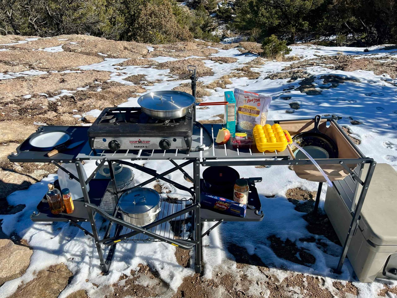 The GCI Master Cook Station stands outside in snow and rocky terrain. Its shelves are full of kitchen items with a sink full of dirty dishes. A cooler is on the ground next to it.