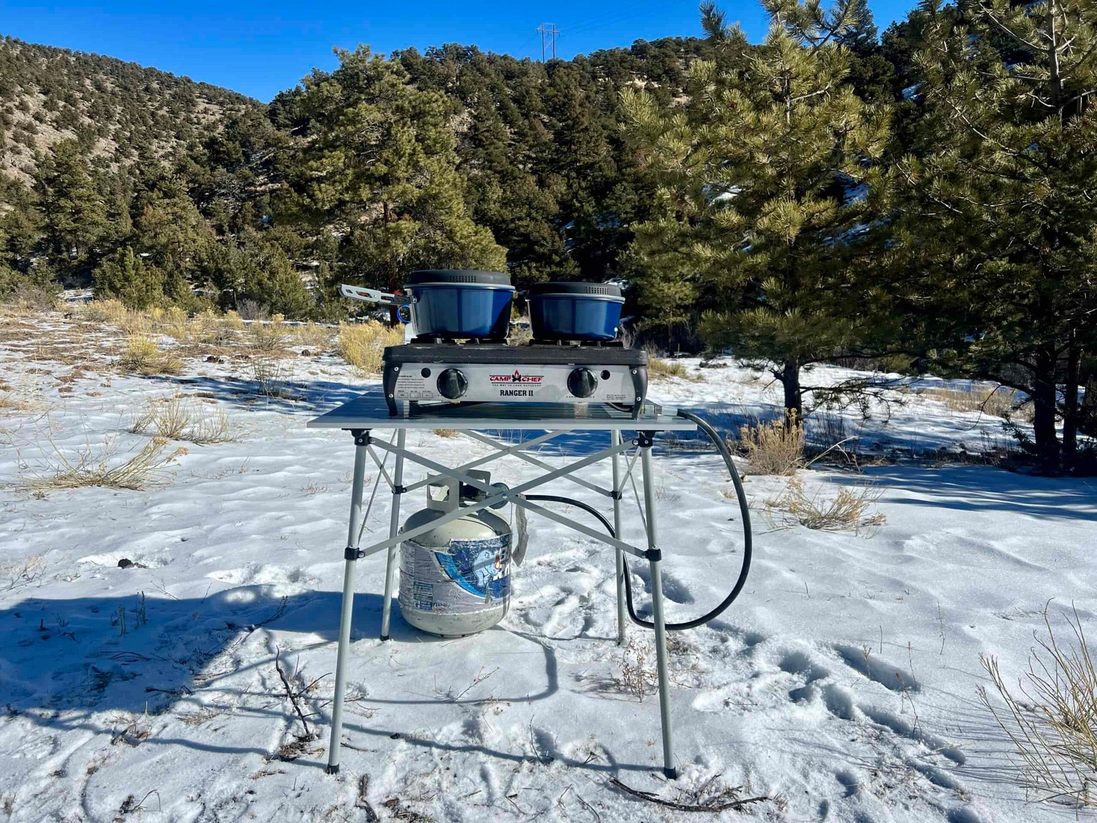 The Coleman Compact Roll-Top Table stands on snow with a two-burner stove and blue cookware on its top. A propane tank sits on the ground behind the table. The background is oak-tree covered hills.