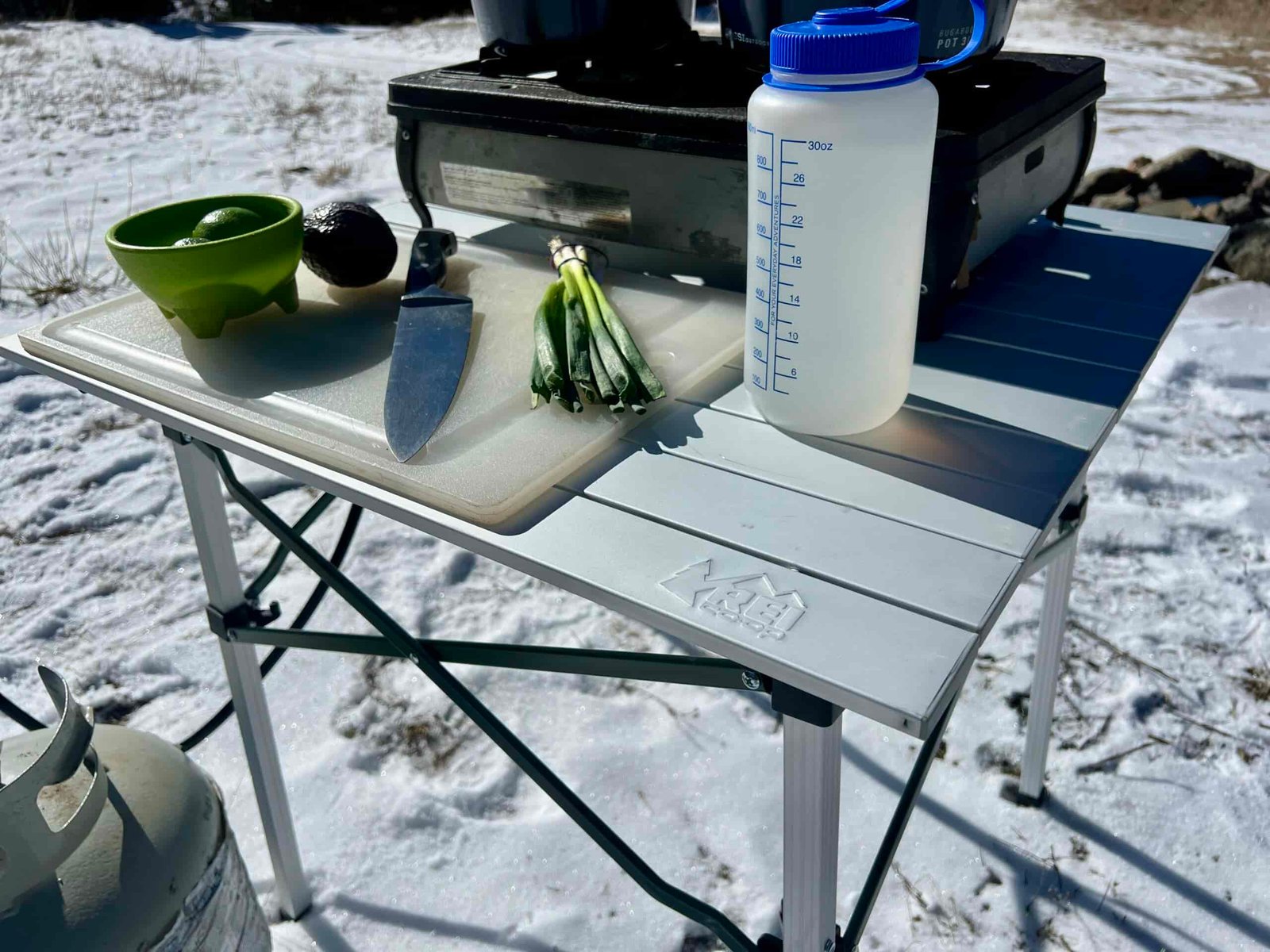 The REI Camp Roll Table stands in the snow. It has a Nalgene, cutting board, knife, scallions, green bowl, limes, and two-burner stove on top.