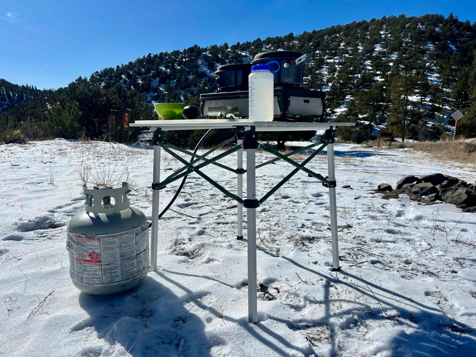 The REI Camp Roll Table stands in the snow with a tree-covered hill in the background. It has a Nalgene, two-burner stove, green bowl, and an avocado on its top. A propane stove sits on the ground next to the table.
