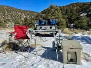 The Portal Roll-Up Table is standing on snow with a stove and cookware on top of it. There is a beige cooler and a red chair next to it. The background is tree-covered hills.