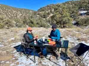 Two people sit and smile around the Mountain Summit Gear Heavy Duty Roll Top Table. They are wearing black and blue jackets and black and orange hats. They have playing cards in their hands. There are bowls, food, and drinks on the table.