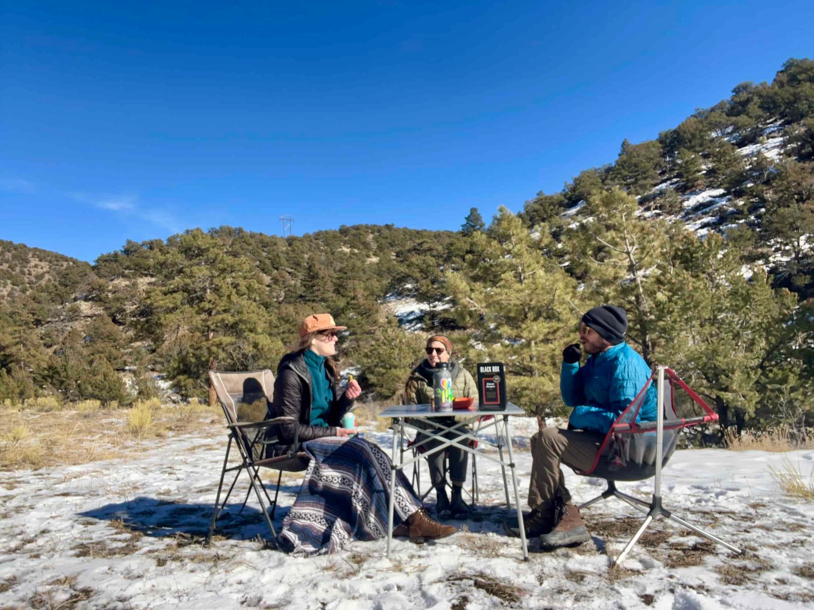 Three people sit around the Coleman Compact Roll-Top Table and are smiling. The background are snow and oak-covered hills. The table has a box of wine and a water bottle.