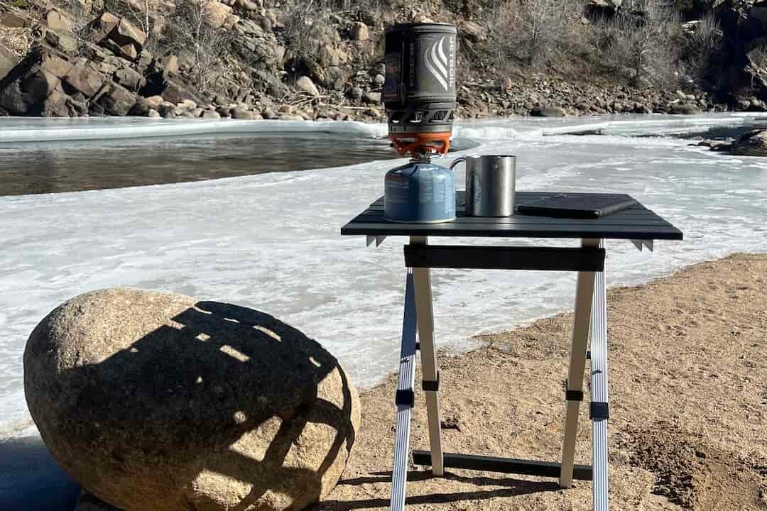 The GCI Compact Table 20 stands in the sand along a river. Ice is covering most of the river. A large boulder is next to the table.