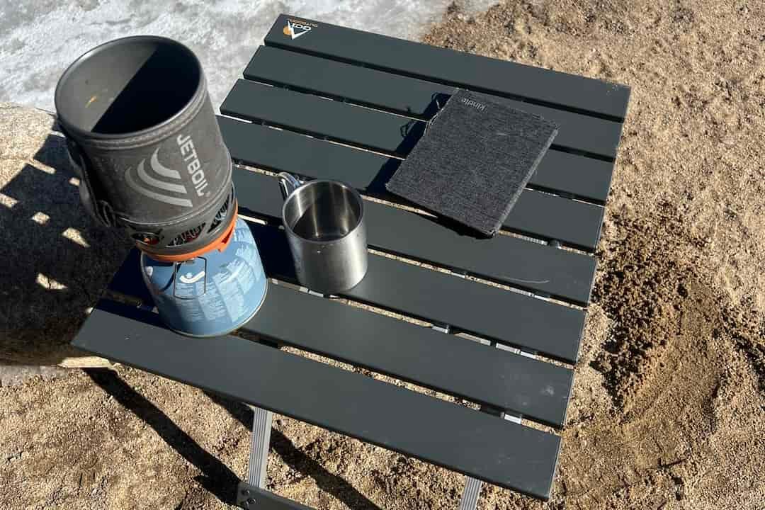 The GCI Compact Table 20 stands on the sand. A jetboil, stainless cup, and kindle sit on top.