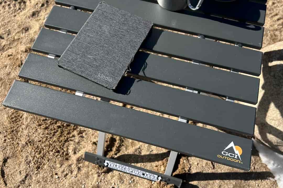 The GCI Compact Table 20 stands in the sand. It has a visible Kindle and stainless cup on it.
