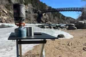 The GCI Compact 20 stands on a sandy river beach. The river is mostly iced over. A Jetboil, stainless cup, and kindle sit on top of the table. In the background, a bridge crosses the river.