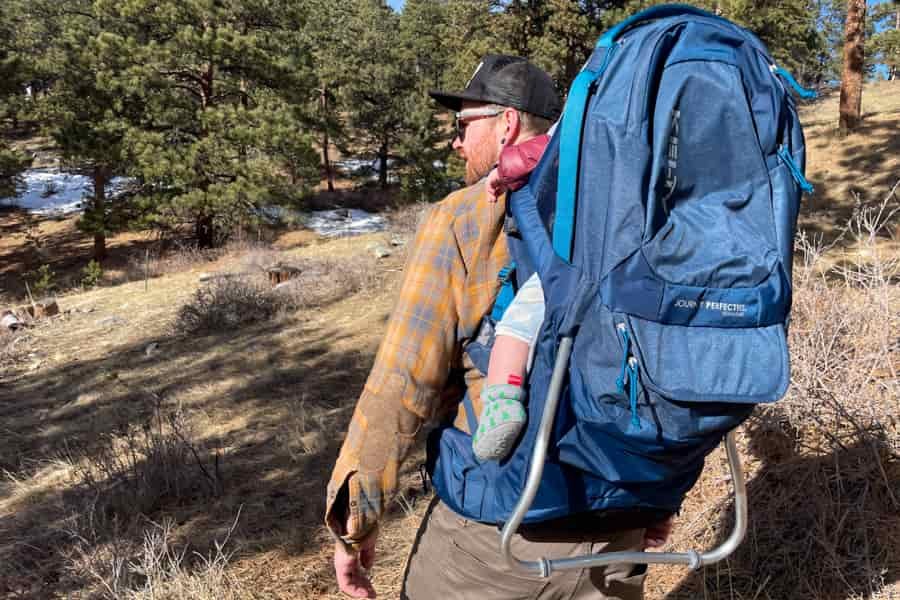 A man hiking through a forested area with the Kelty PerfectFIT Journey Signature carrier, showcasing its ergonomic design and sturdy kickstand.