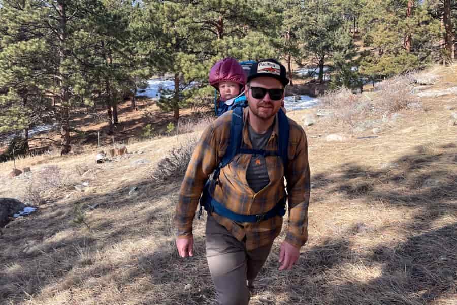 A front-facing shot of a man hiking uphill, wearing the Kelty PerfectFIT Journey Signature carrier with a child seated comfortably.