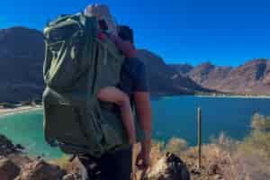 A scenic view of a person hiking along a rocky path overlooking a stunning lake and mountains, with the Osprey Poco Premium child carrier in green, carrying a child.