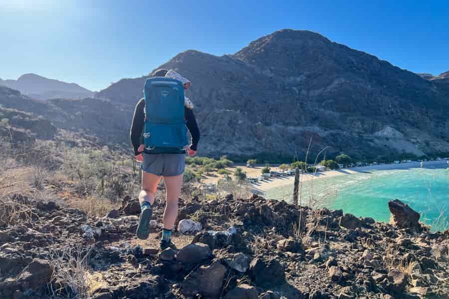 Person hiking down a rocky trail with the Osprey Poco SLT carrier, capturing the contrast of the bright blue carrier against the rocky landscape.