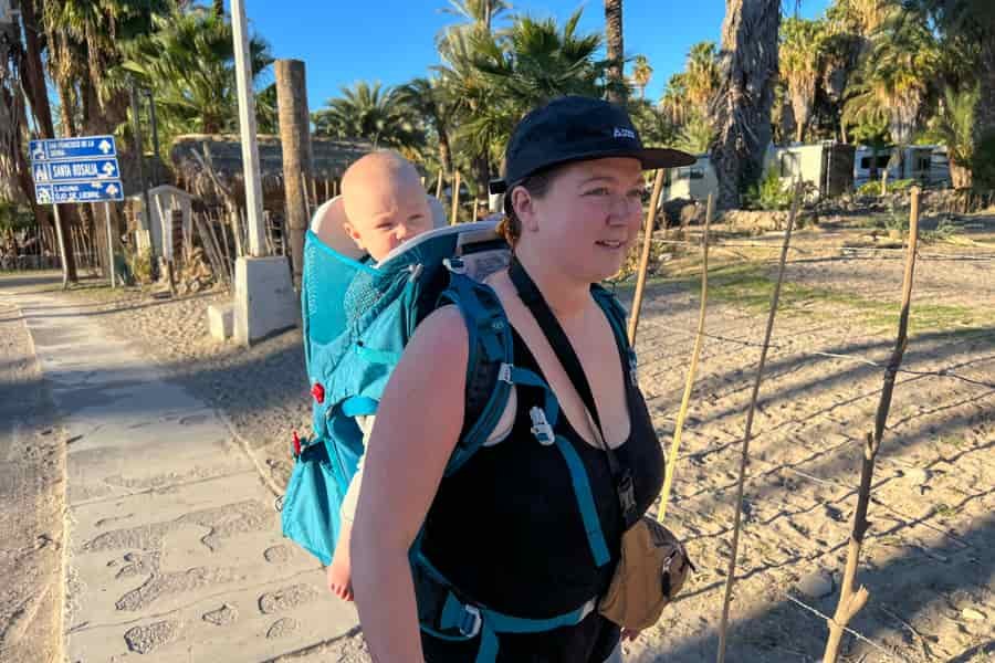 Woman carrying baby in a blue Osprey Poco SLT child carrier, walking along a sandy path with palm trees in the background.