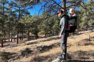 A parent wearing the Thule Sapling child carrier with a toddler, standing in a scenic forest area under a sunny sky.