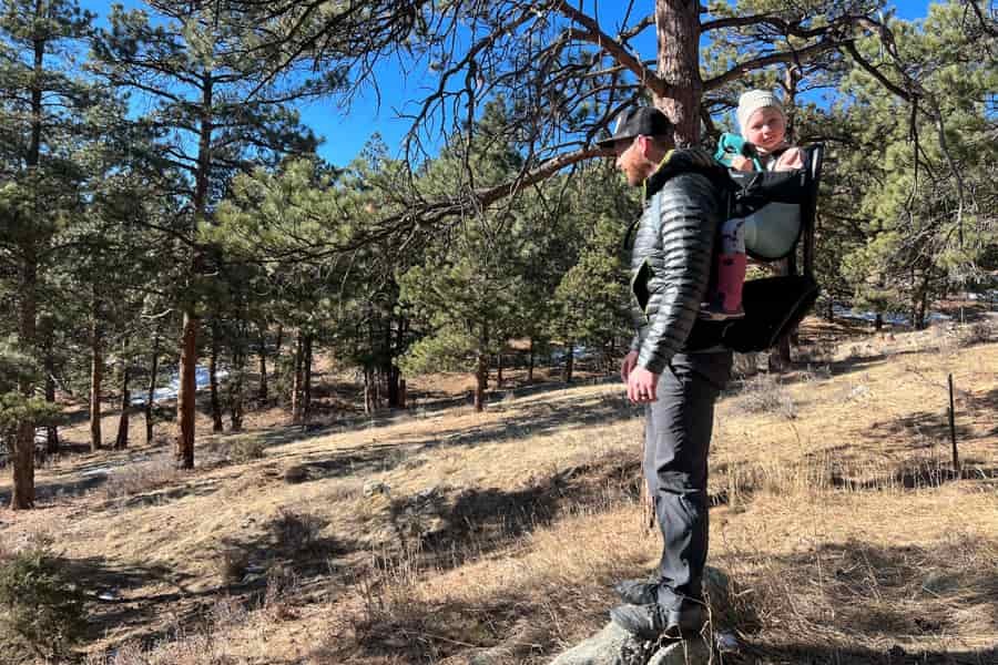 A parent wearing the Thule Sapling child carrier with a toddler, standing in a scenic forest area under a sunny sky.
