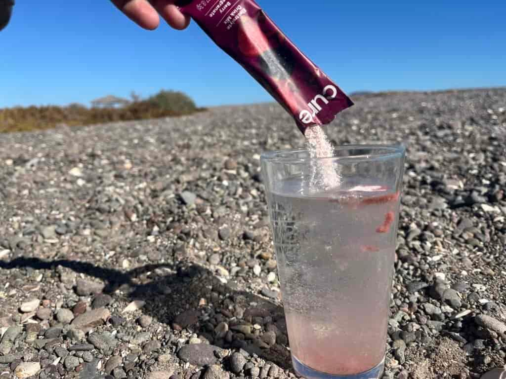 A packet of Cure electrolytes being poured into a glass on a rocky beach