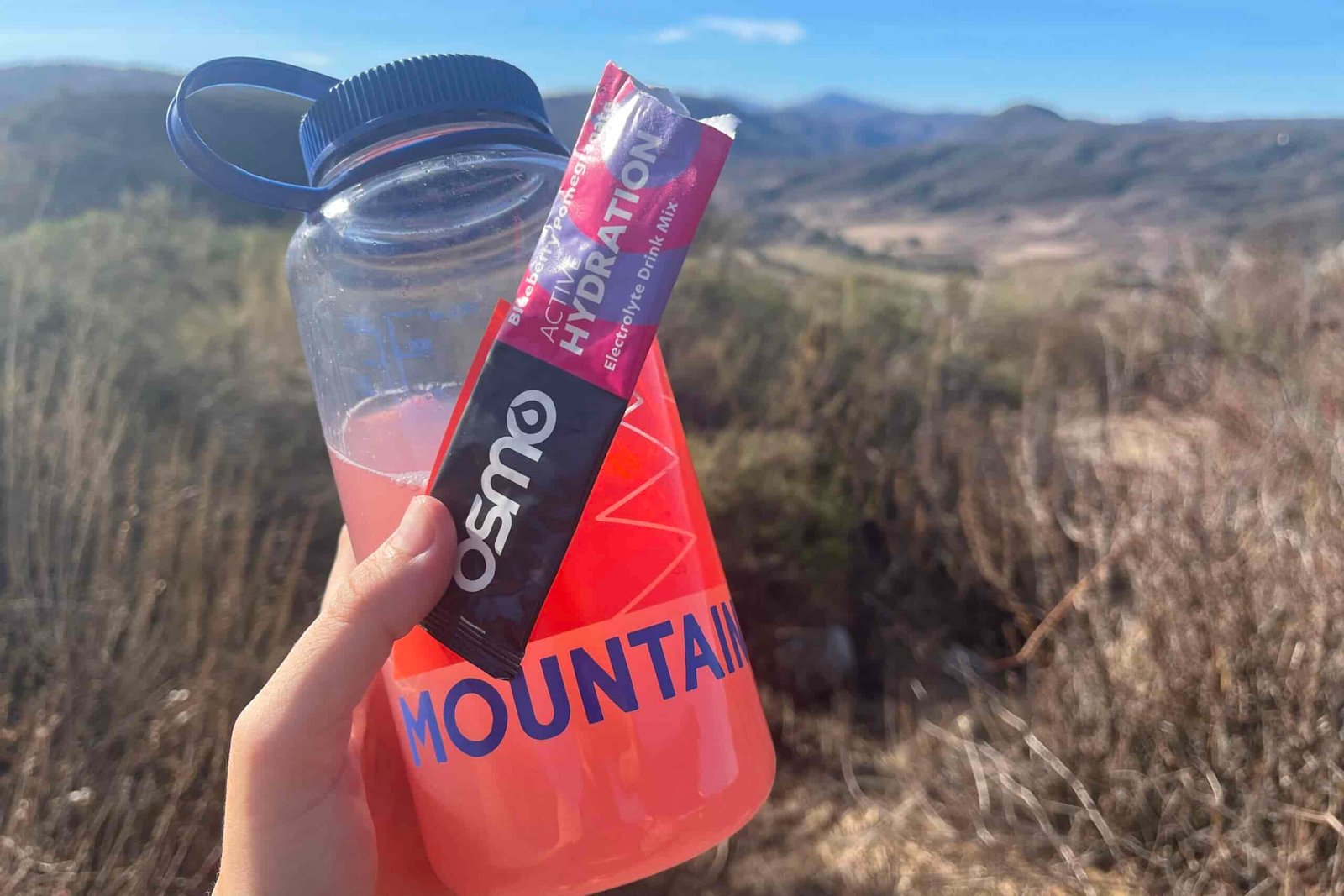 A hand holding up Osmo Active Hydration powder mixed with water in a Nalgene bottle, with mountains in background