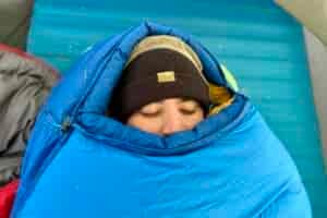 Close-up image of a person's face barely peeking out of a closed mummy bag with the mummy hood up and the bag closed to the top.