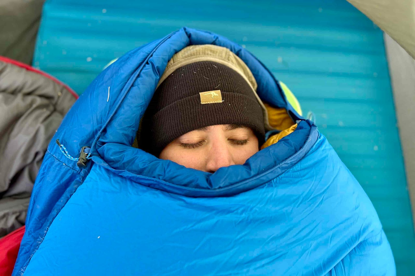 Close-up image of a person's face barely peeking out of a closed mummy bag with the mummy hood up and the bag closed to the top.