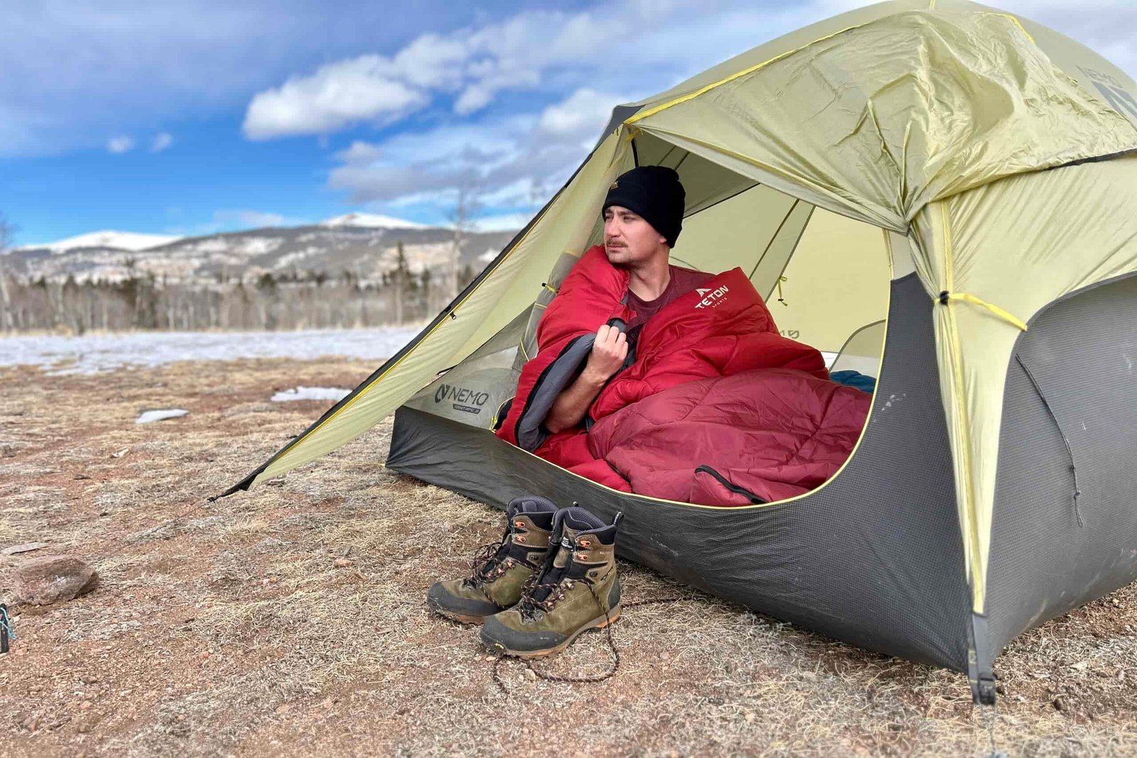 A man looks out of a tent into a snowy, mountainous landscape with a sleeping bag wrapped over his shoulders.
