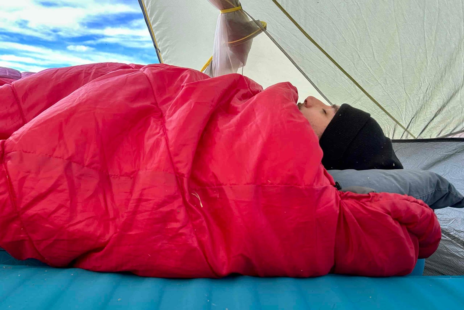 Close-up side profile image of a person sleeping in a big sleeping bag in a tent. Their head is just barely visible poking out.