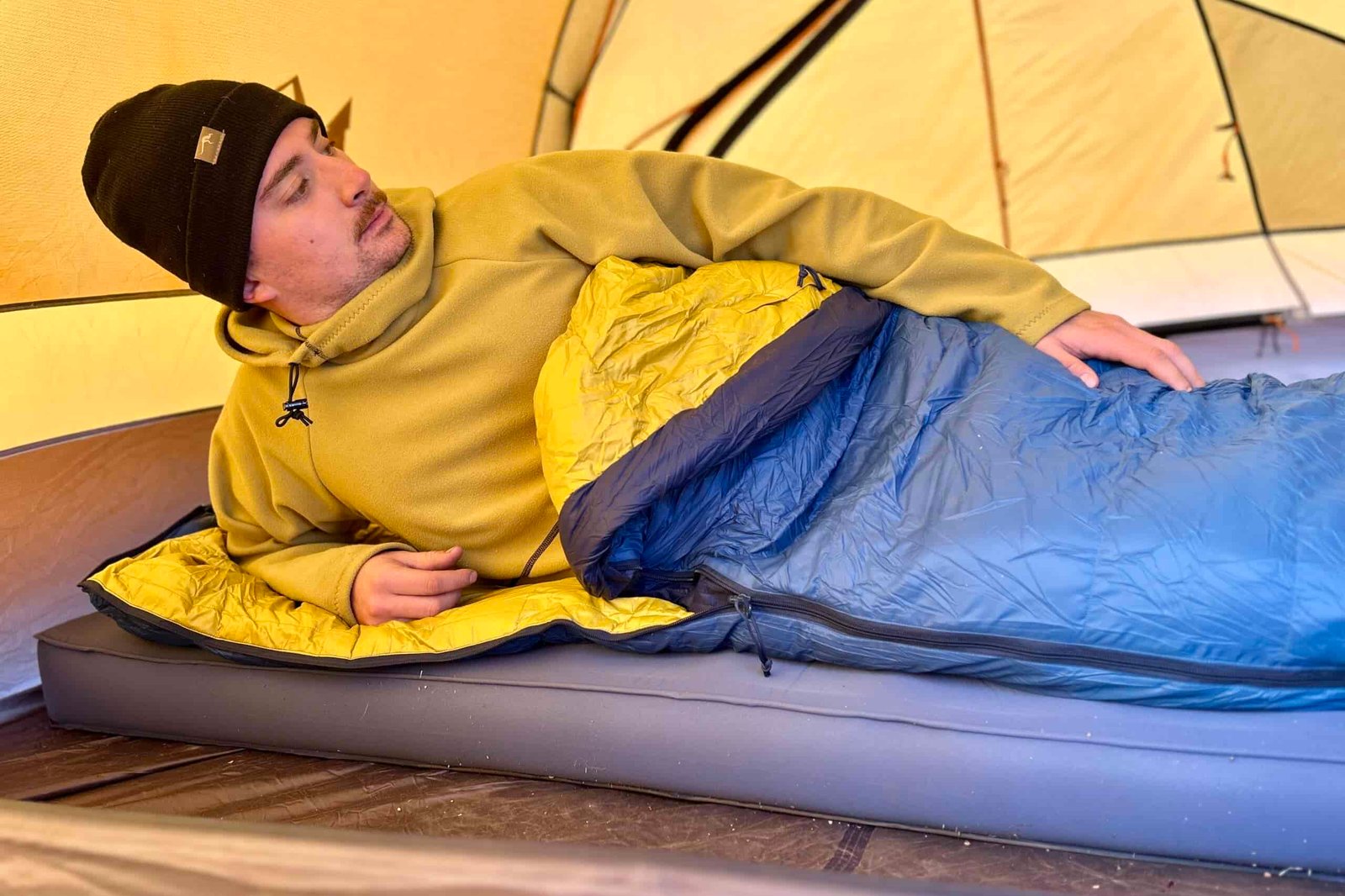 A man sits up on one arm in a a sleeping bag on a camping mattress in a tent.