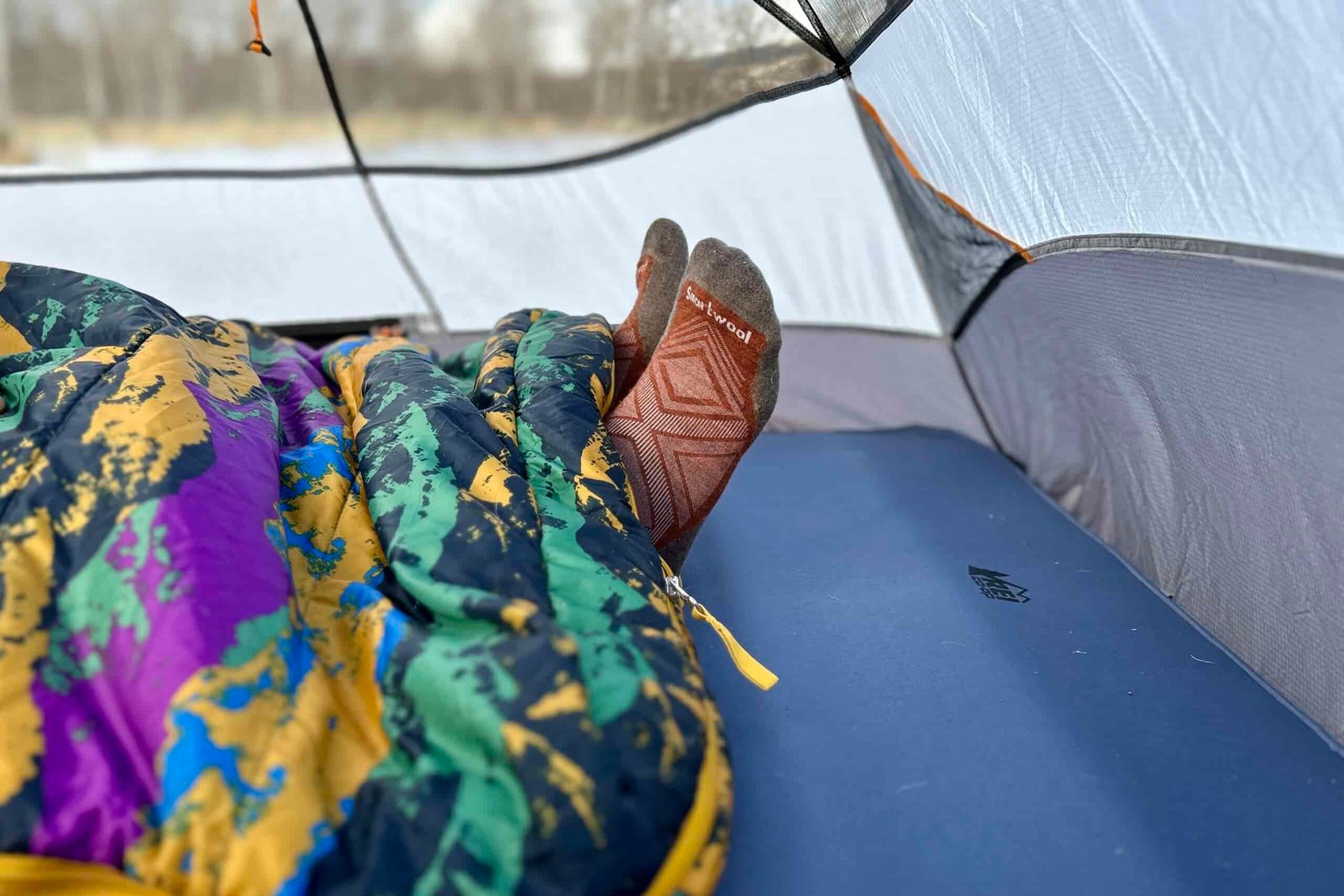 Close up image of the bottom of a sleeping bag with a person's feet sticking out and the zipper pull visible.
