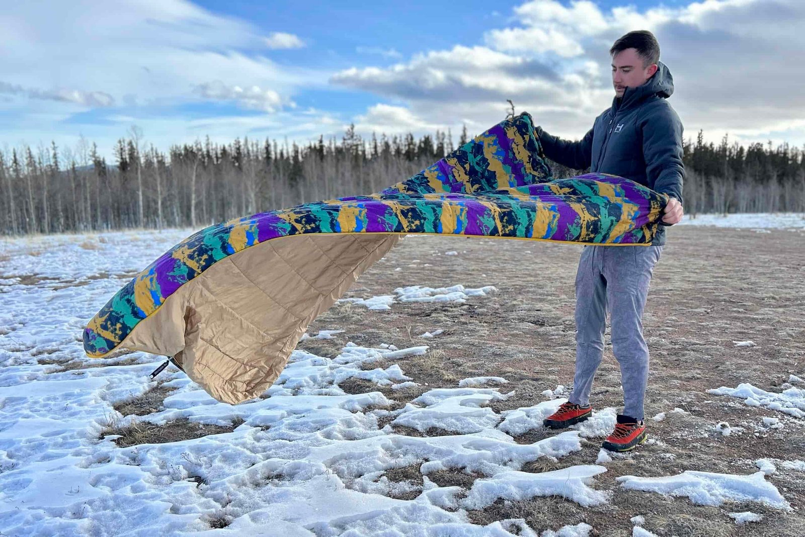 A man in a snowy forested landscape fluffs out a sleeping bag.