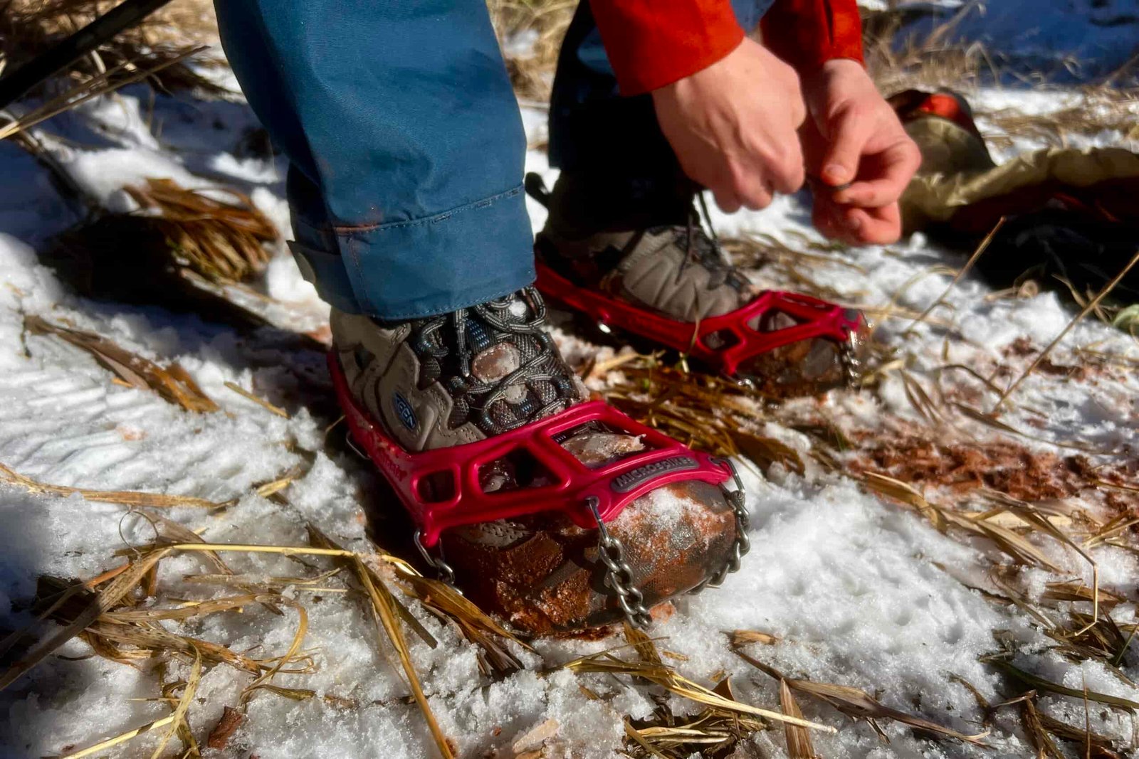 A hiker puts on a pair of ice cleats against a snowy ground.