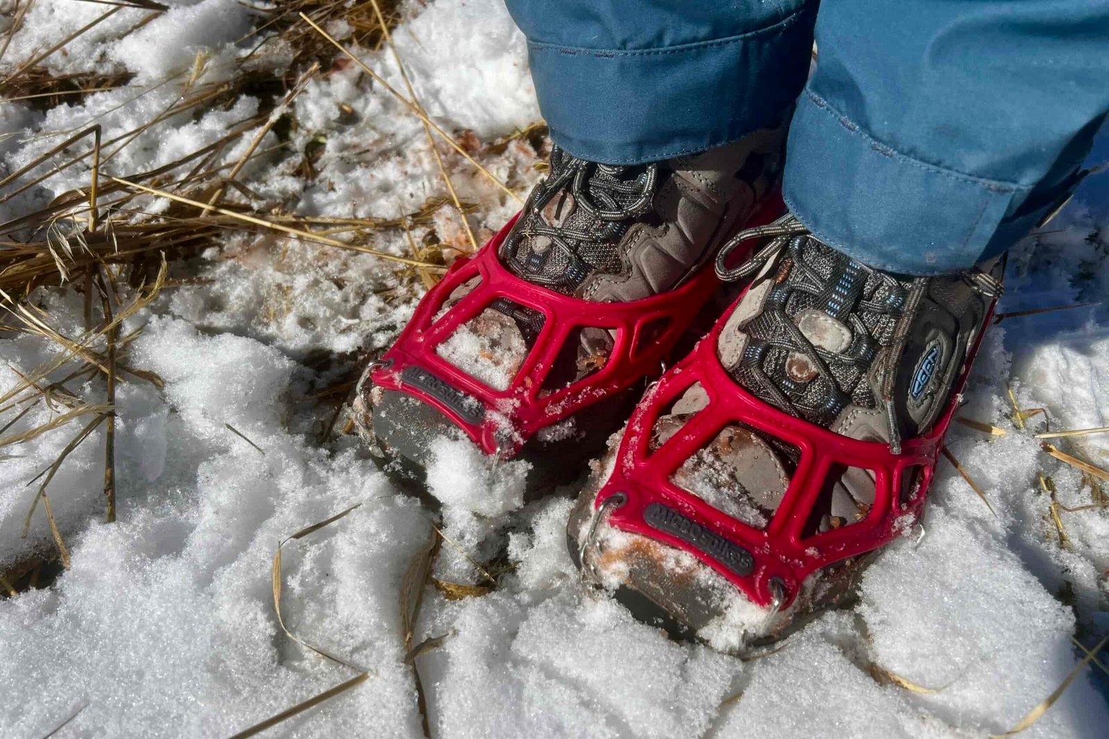 A top down view of feet with hiking shoes wearing a pair of ice cleats on snowy ground.