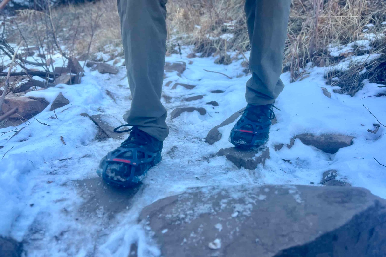 A hiker's feet walk over ice and rocks with a pair of ice cleats.