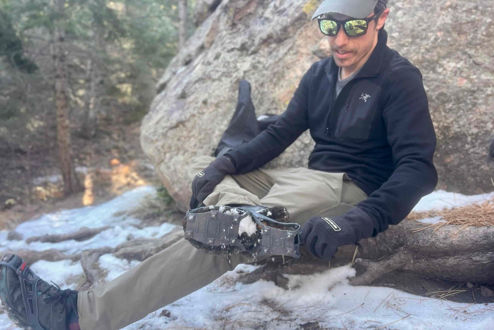 A hiker sits on a rock while pulling a pair of ice cleats onto his feet.