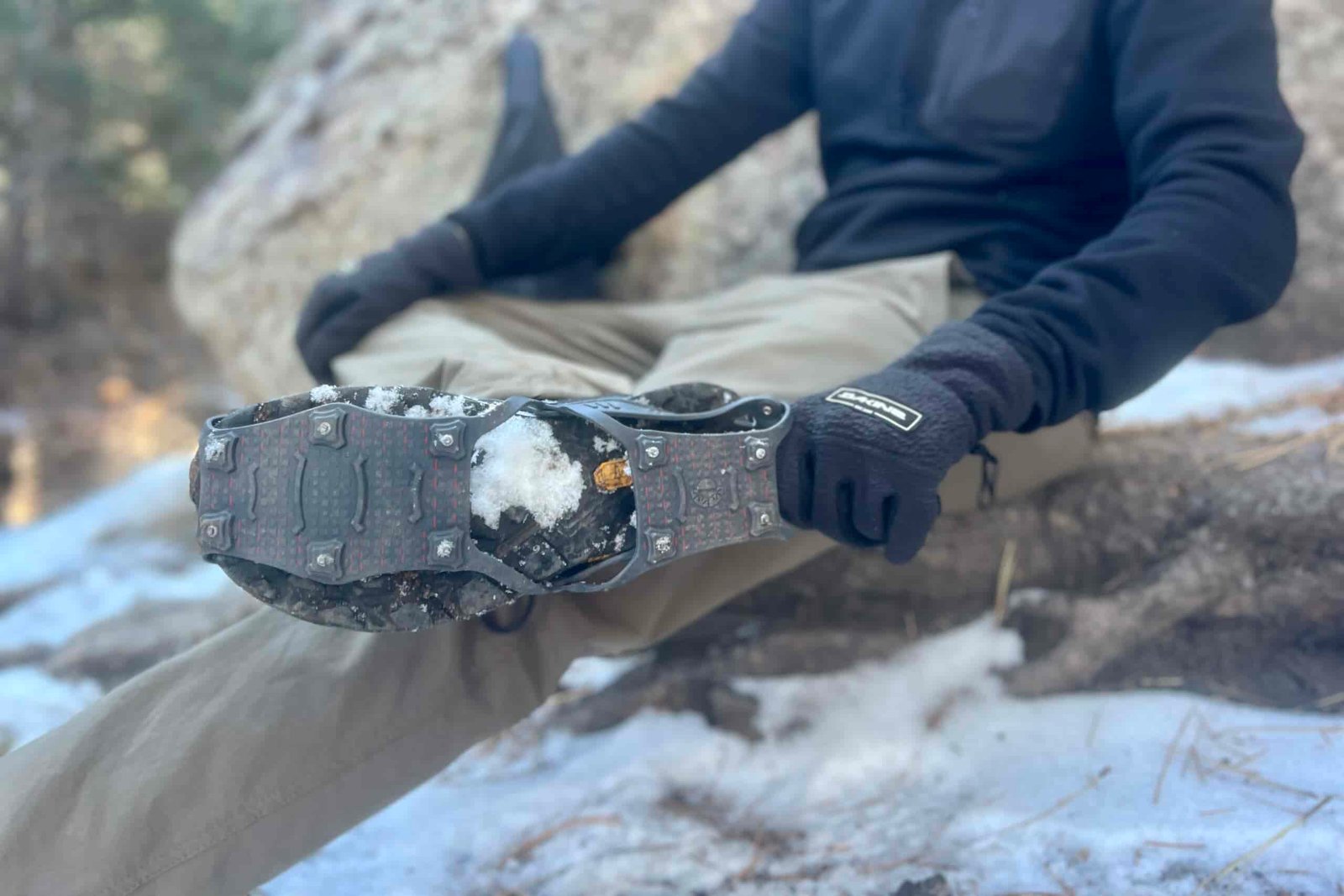 A hiker sits on a rock while pulling a pair of ice cleats onto his feet.