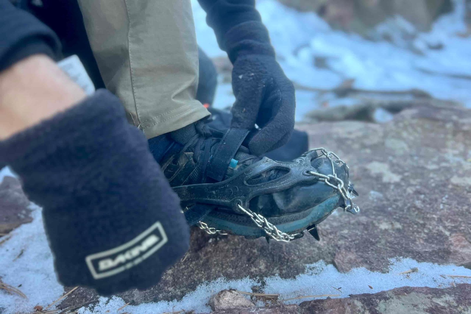 A hiker pulls on a pair of ice spikes against a snowy, rocky background.