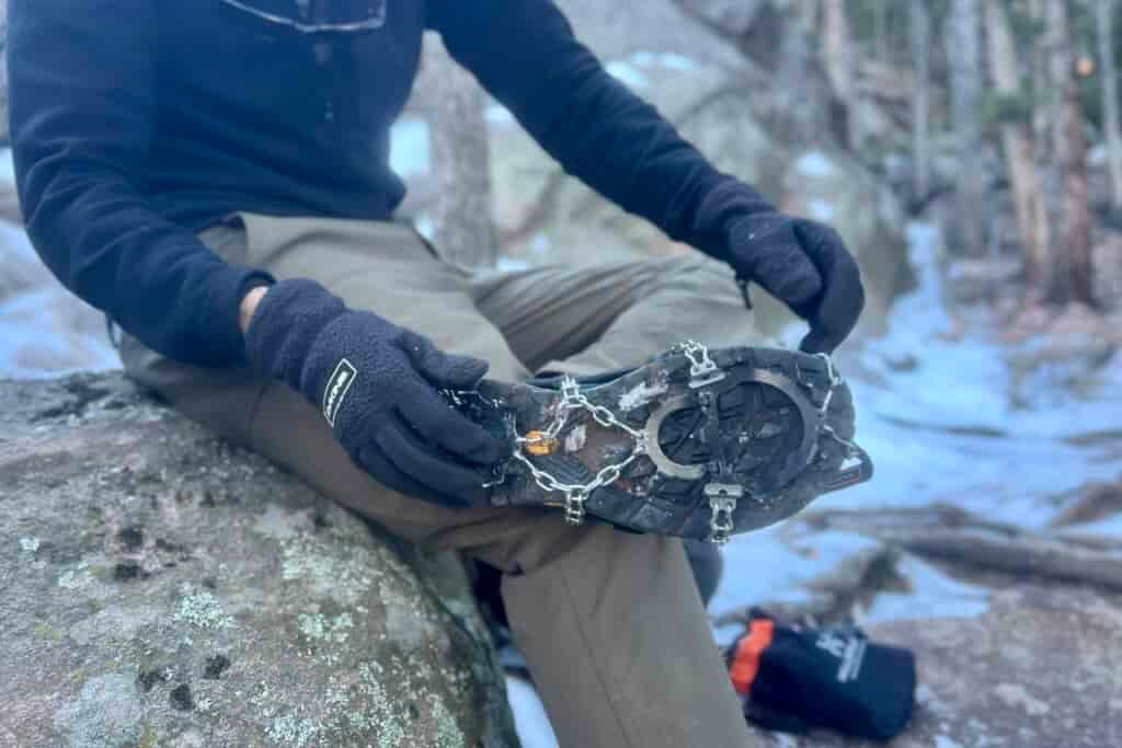 A hiker pulls on a pair of ice spikes against a snowy, rocky background.