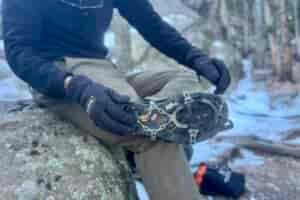 A hiker pulls on a pair of ice spikes against a snowy, rocky background.