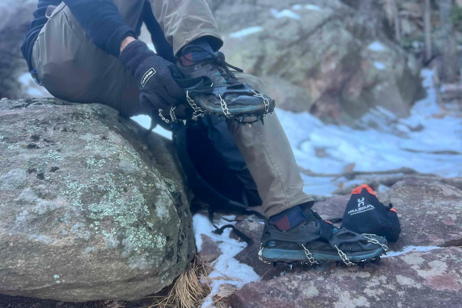A hiker pulls on a pair of ice spikes against a snowy, rocky background.