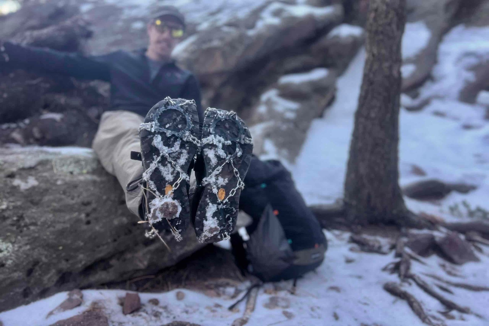 A hiker sits on a rock with his feet stretched out towards the camera. He is wearing ice spikes on his feet. Snowy forest background.