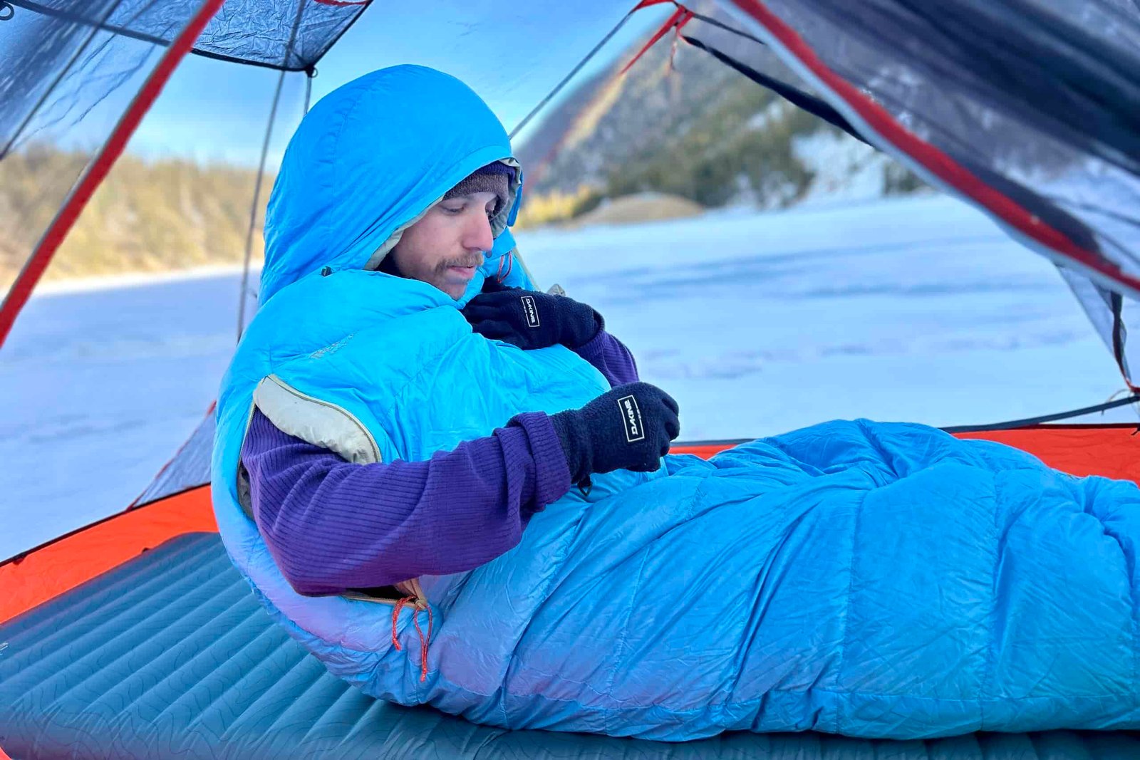 Close up image of a man sitting up in a sleeping bag with his arms sticking out of arm holes and the hood up over his head.