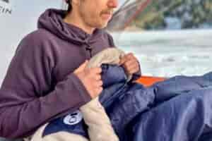 Close up image of a man sitting up in a sleeping bag in a tent in a wintry setting with their hands on the small draft tube at the collar.