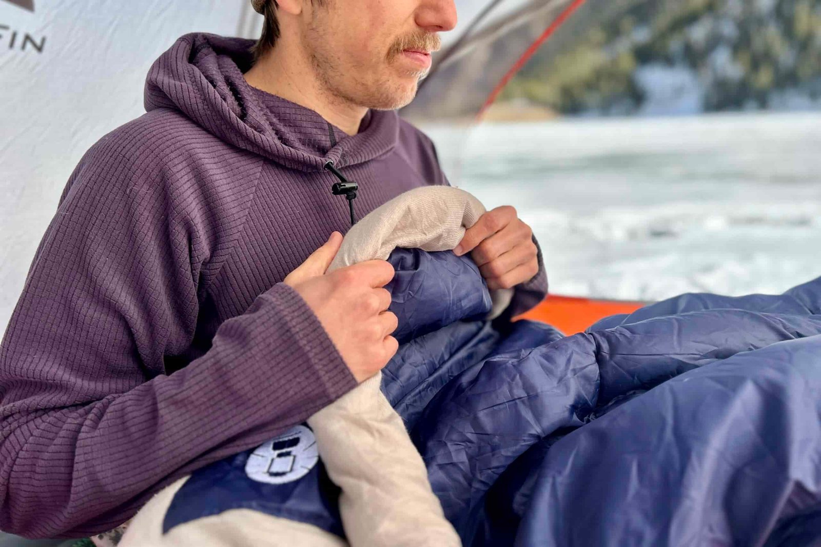 Close up image of a man sitting up in a sleeping bag in a tent in a wintry setting with their hands on the small draft tube at the collar.