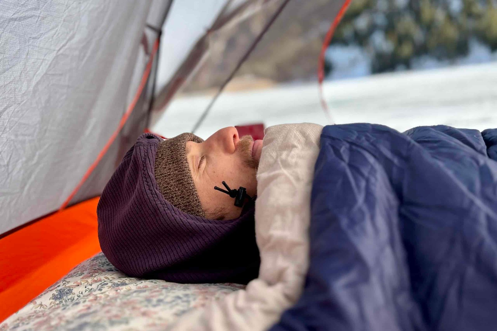 Side-profile of a man sleeping in a sleeping bag with the bag pulled up to his chin, and a hood and hat on.