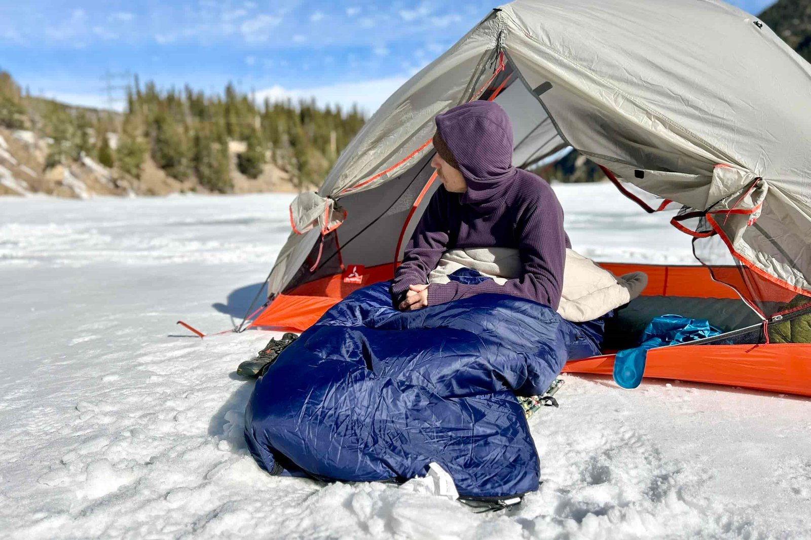 A man in a snowy field sits in a tent wrapped in a sleeping bag looking off in the distance.