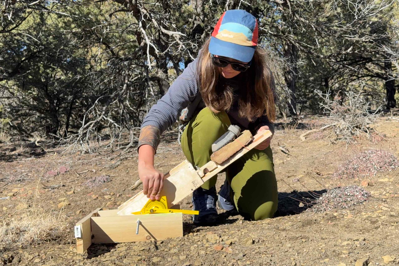 A person is testing boot traction on the Ruffwear boots. They are holding up a wooden board with a rock on top. The boot is on top of the rock. They are also holding a protractor to measure the angle of the board. The test is being performed in a pinion forest.