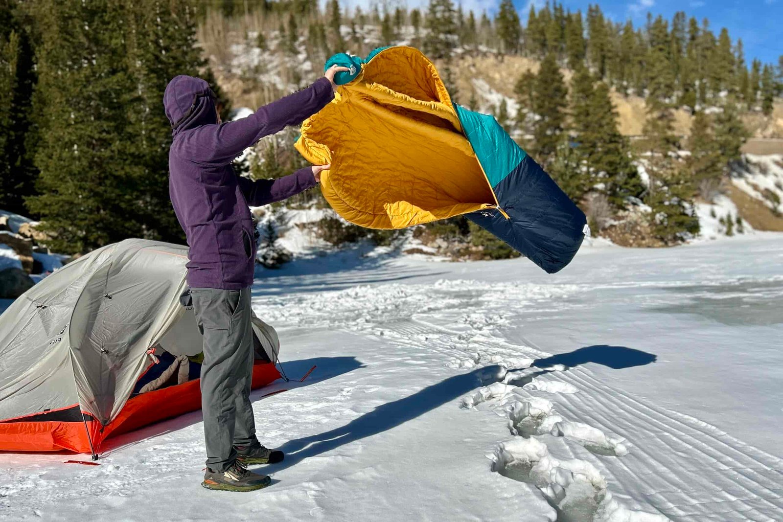 A person fluffs out a sleeping bag in a wintry setting next to a tent.
