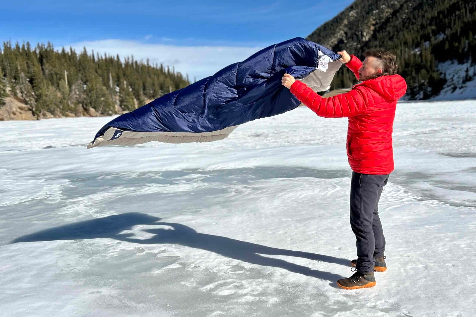 A man fluffs a sleeping bag in a snowy mountain setting.